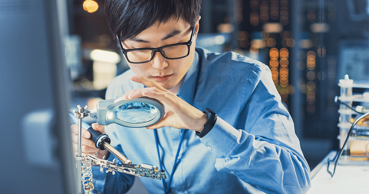 An engineer working in a laboratory solders a circuit board while looking through a magnifying glass.