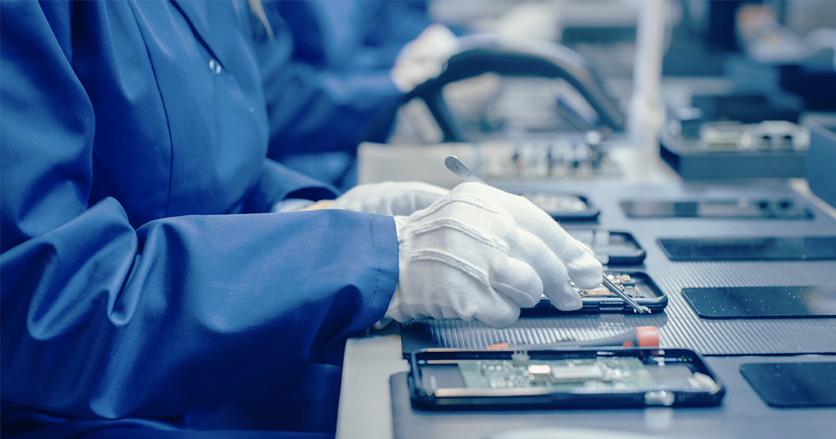 Three factory workers wearing blue lab coats and protective eyewear assembling smartphone PCBs | Sourcengine