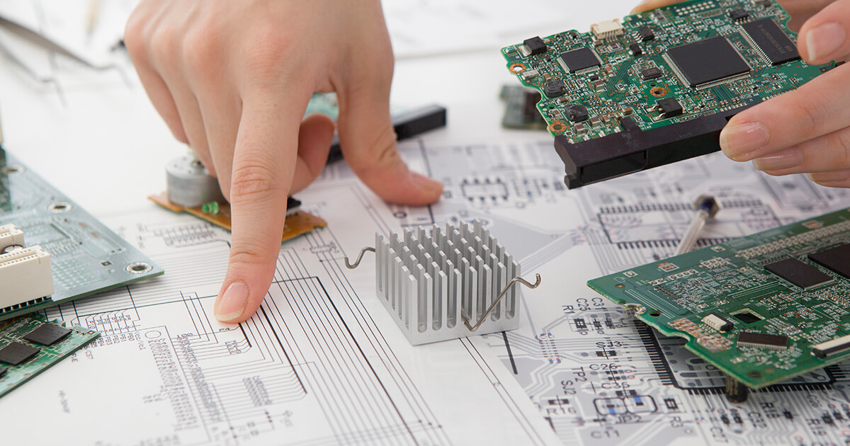 An engineer holding a fully assembled PCB exams microchip schematics on a table covered with chipsets.