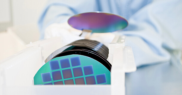 An engineer holds a silicon wafer with a pair of tongs in front of a clear plastic case holding a series of silicon wafers. | Sourcengine 