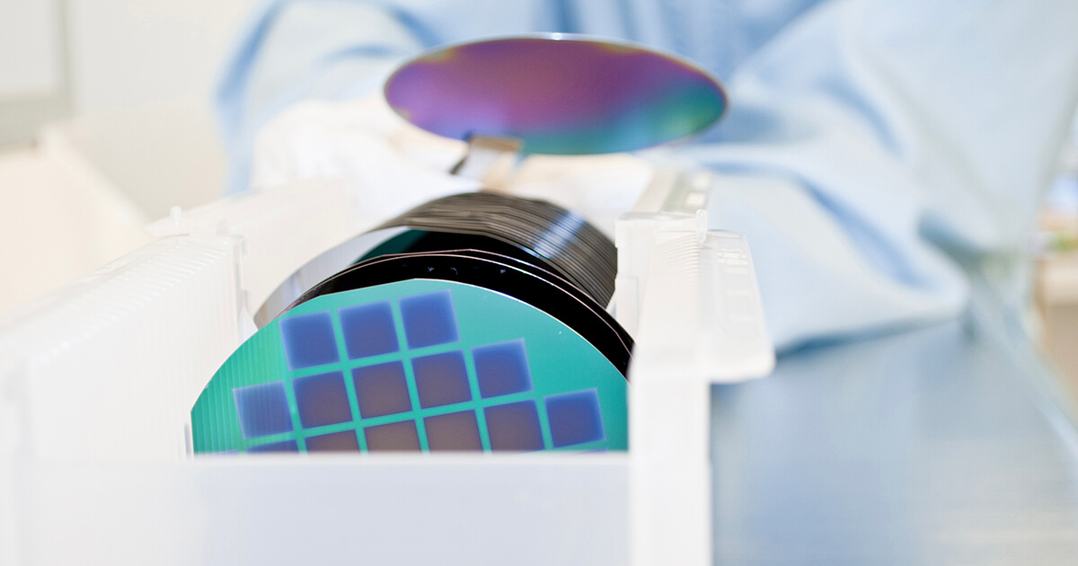 An engineer holds a silicon wafer with a pair of tongs in front of a clear plastic case holding a series of silicon wafers. | Sourcengine 