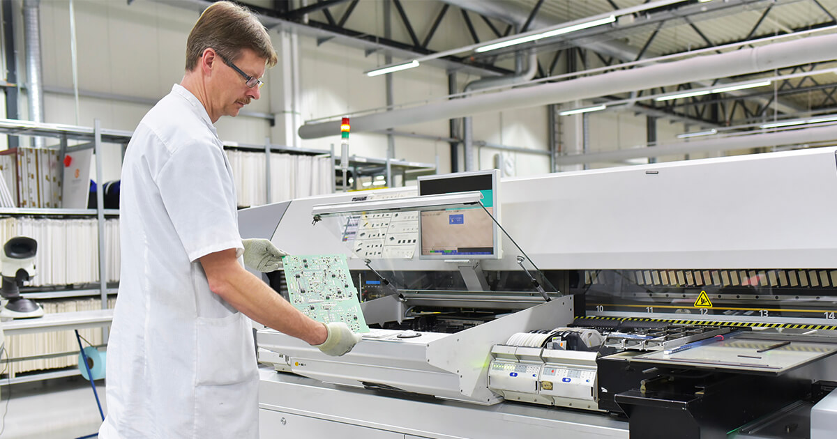 A man in a white coat holding a fully assembled PCB stands in front of a piece of manufacturing equipment. | Sourcengine