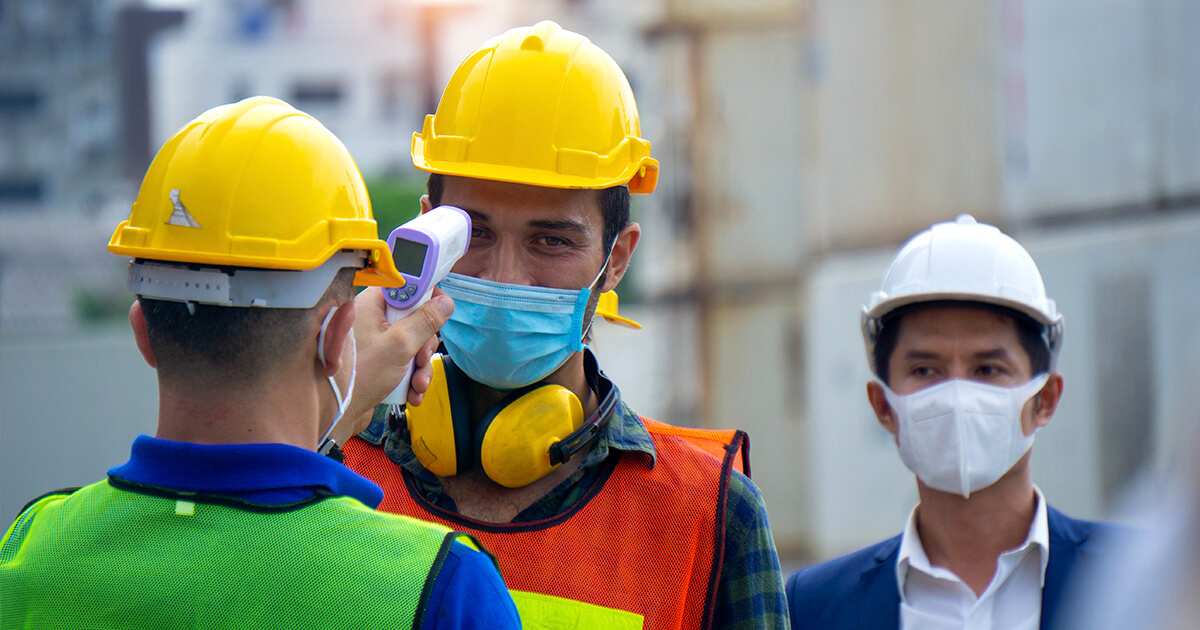 A worker performs a temperature check on another worker while a third observes.