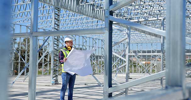 A construction worker holding a blueprint stands inside a metal building frame. 