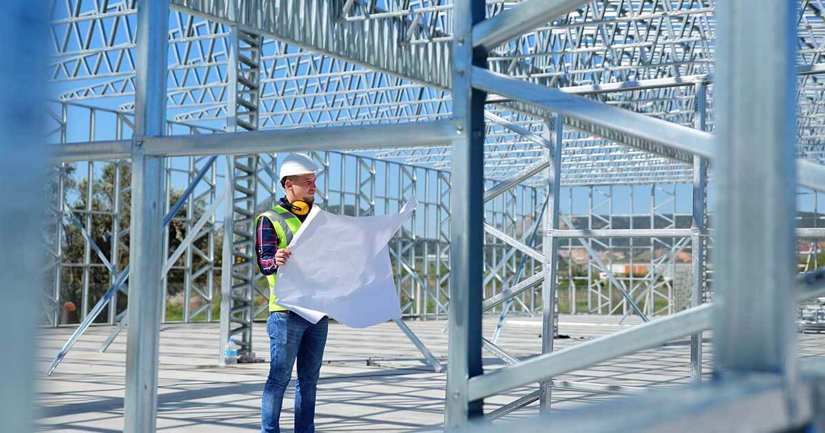A construction worker holding a blueprint stands inside a metal building frame.
