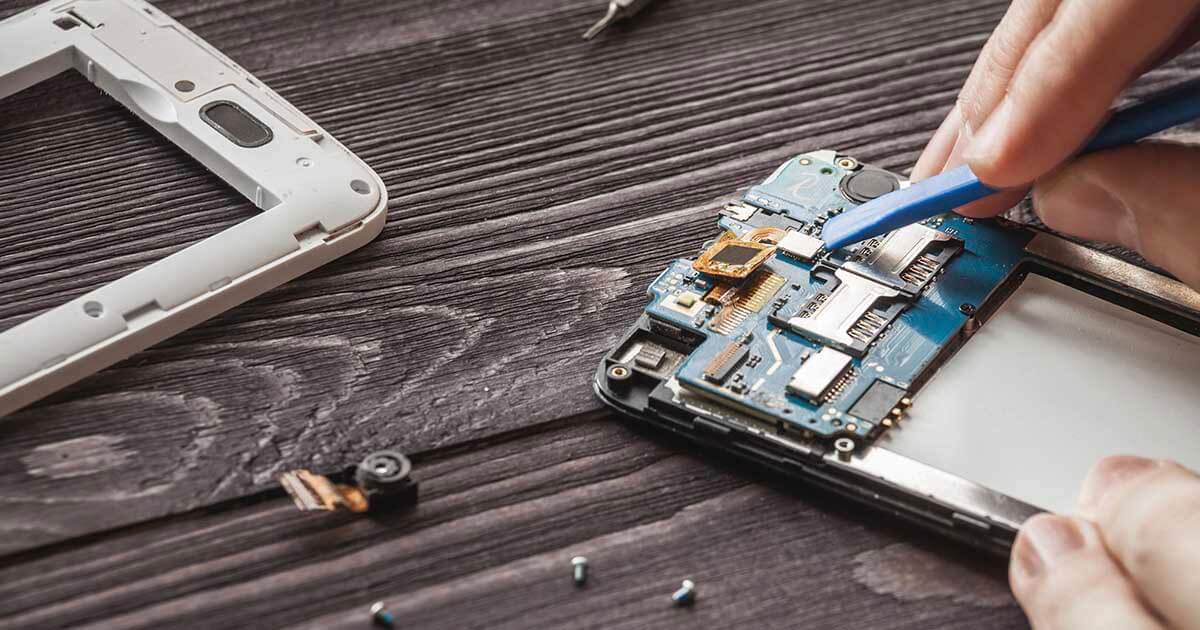 The hands of a technician in the process of carefully disassembling a smartphone using a plastic pry tool.