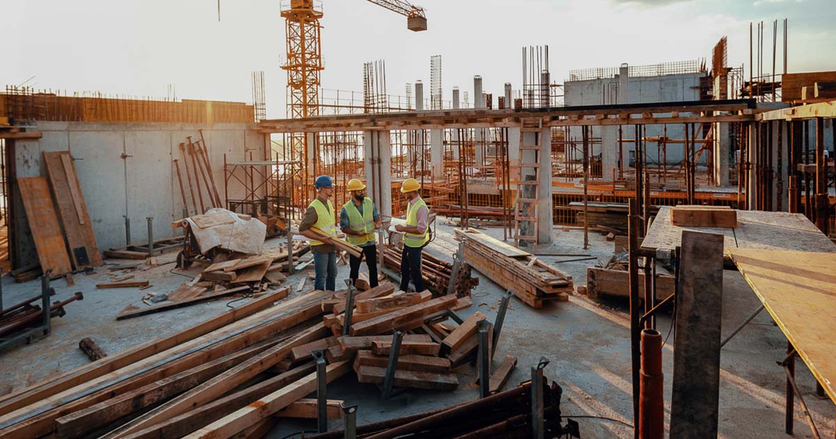 Three construction workers stand in the middle of a large construction site filled with stacks of lumbar, rebar, and AC plywood. 