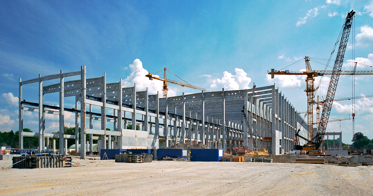 A wide shot of a large construction site that features a massive steel building framework and three tower cranes. 