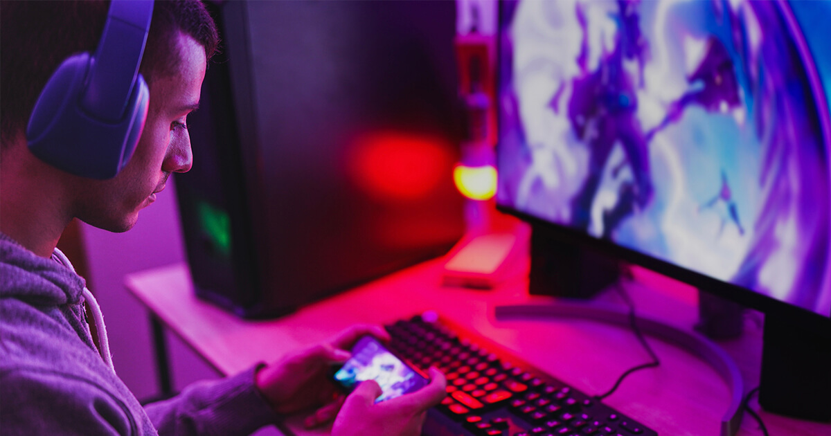 A gamer wearing headphones sitting in front of a high-performance computing rig uses a mobile device as a controller.