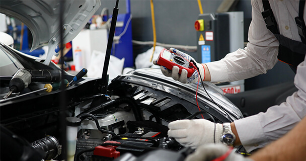 A worker stands in front of a car with its hood open using a voltmeter to check its electrical systems.