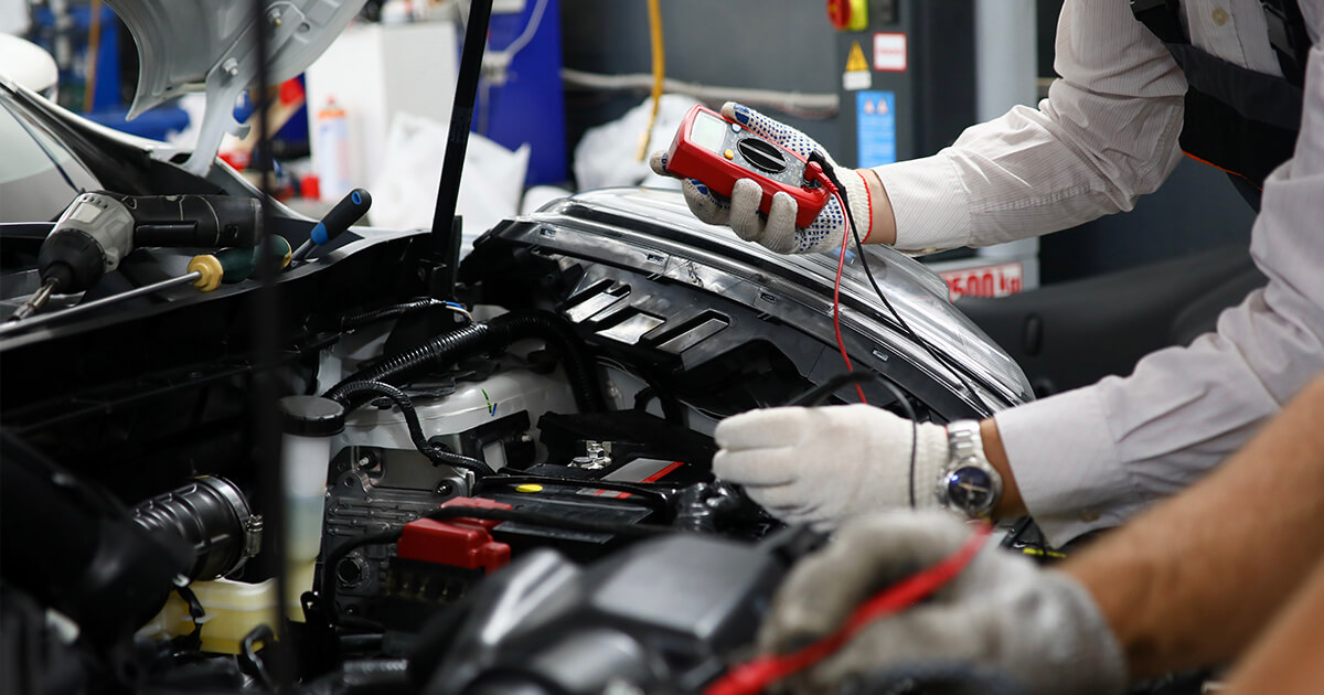 A worker stands in front of a car with its hood open using a voltmeter to check its electrical systems.