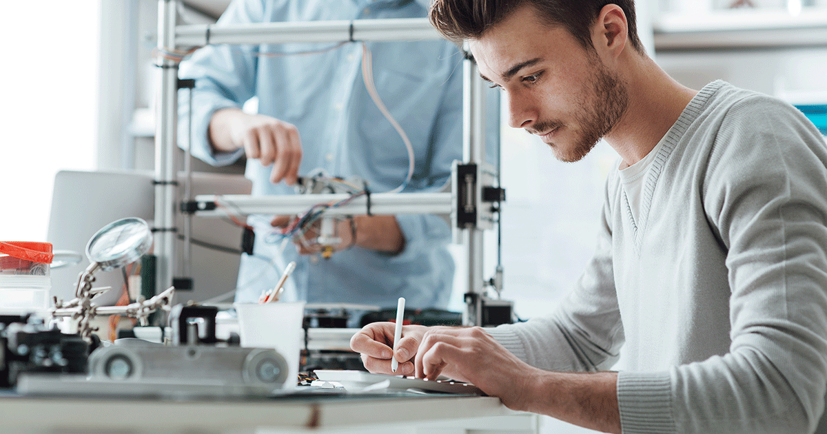A college-age engineering student takes notes at a desk using a tablet computer using a stylus. In the background, another student works on a 3D printer.