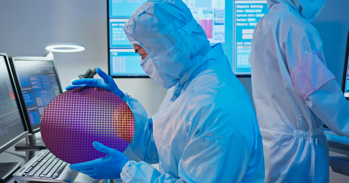 A technician in full personal protective equipment holds a silicon wafer while standing inside a laboratory