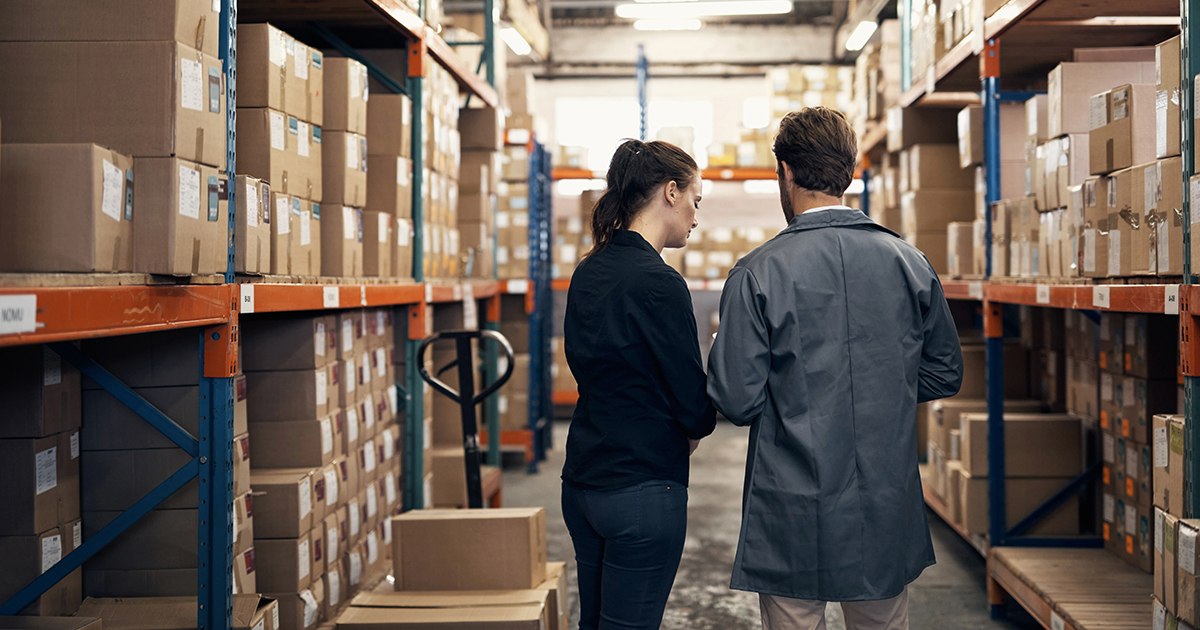 An image of two people walking down a row of stored stock in a warehouse 